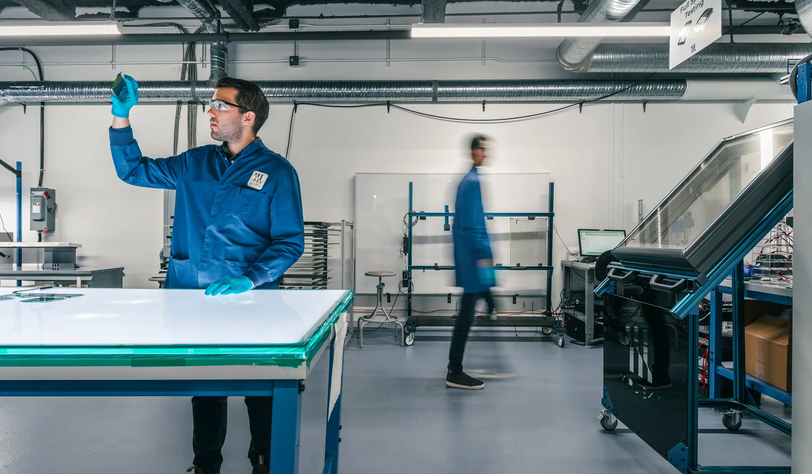 Miru employee in goggles, lab coat, and gloves looking through a small piece of glass; large panes of glass and another employee surround him in a lab.