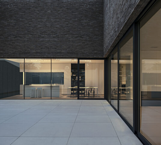 The brightly lit kitchen of a modern home shown through wall-to-wall windows at night.