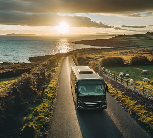 Private bus with large windshield and sunroof alongside sheep on a winding coastal road with the sun setting behind it.