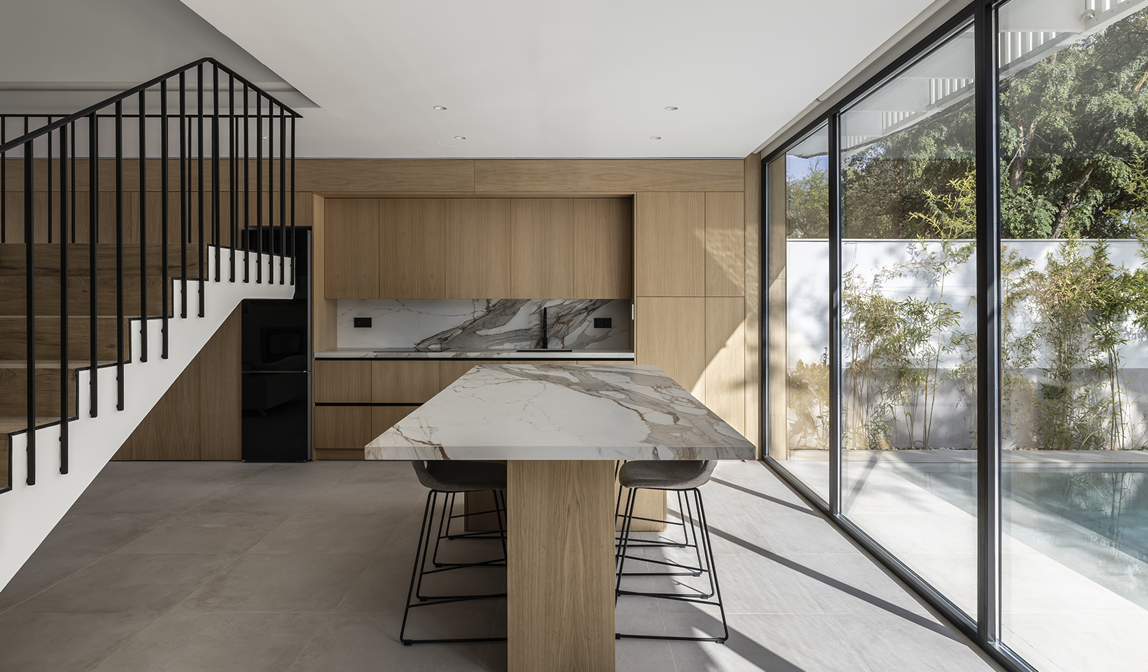 A room with a marble-topped seating area, tall stools, and light wood cabinetry. Exterior with pool, bamboo, trees, and sky shown through clear wall-to-wall windows.