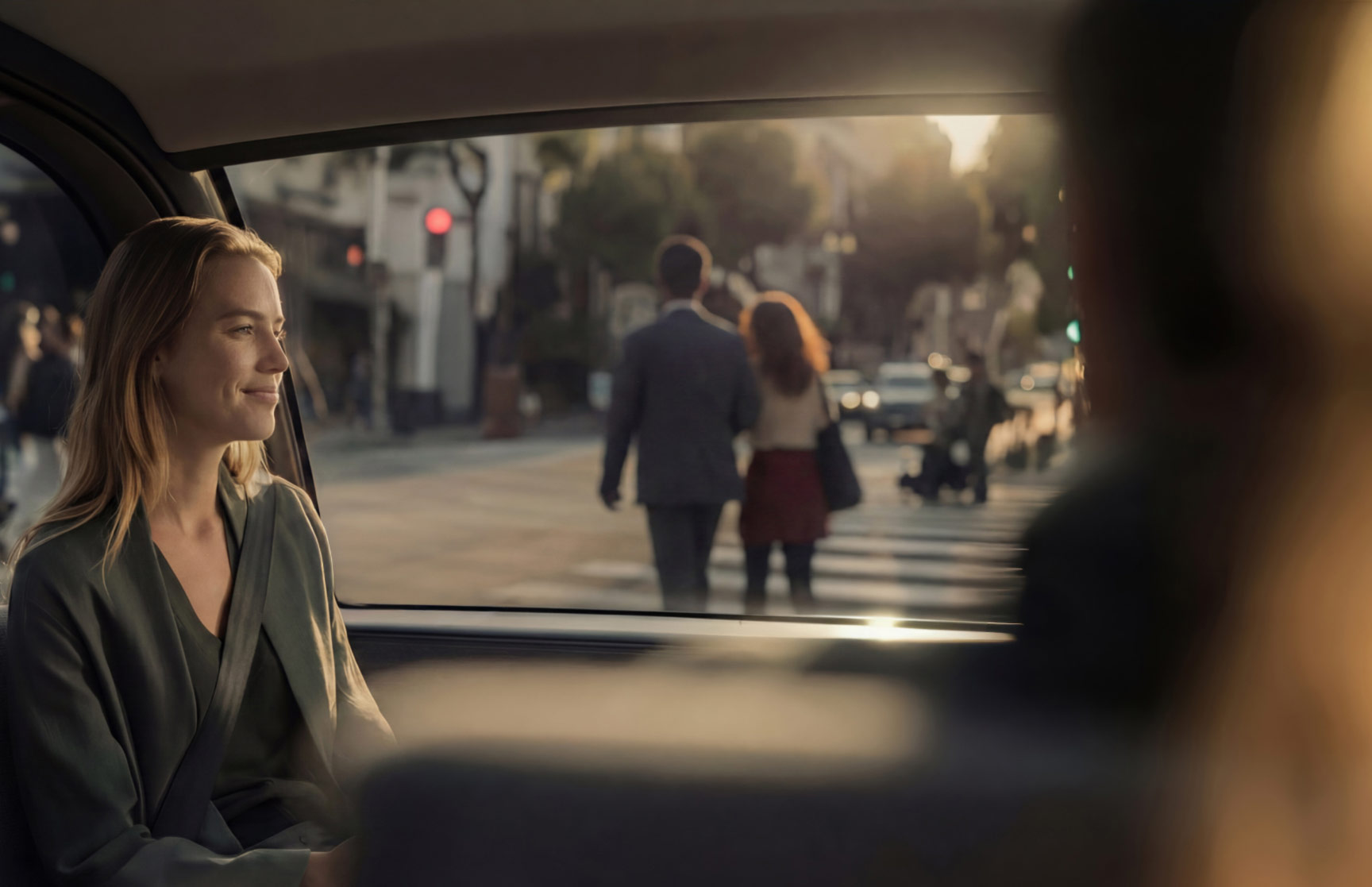 Woman inside an autonomous vehicle; a couple walks in a crosswalk outside. The setting sun appears bright but the interior of the cabin is comfortably dimmed.
