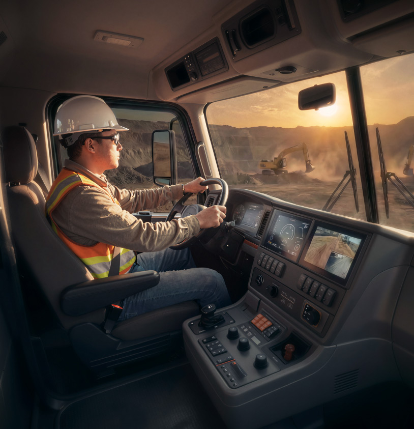 A man in a hard hat drives a vehicle; the setting sun is visible thorugh the tinted windshield.