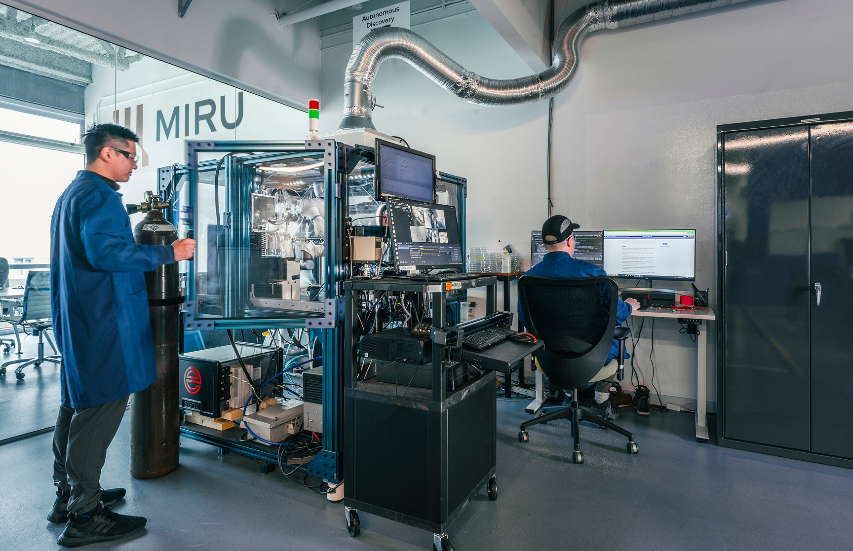 Two Miru employees in blue jumpsuits operate equipment in a laboratory.